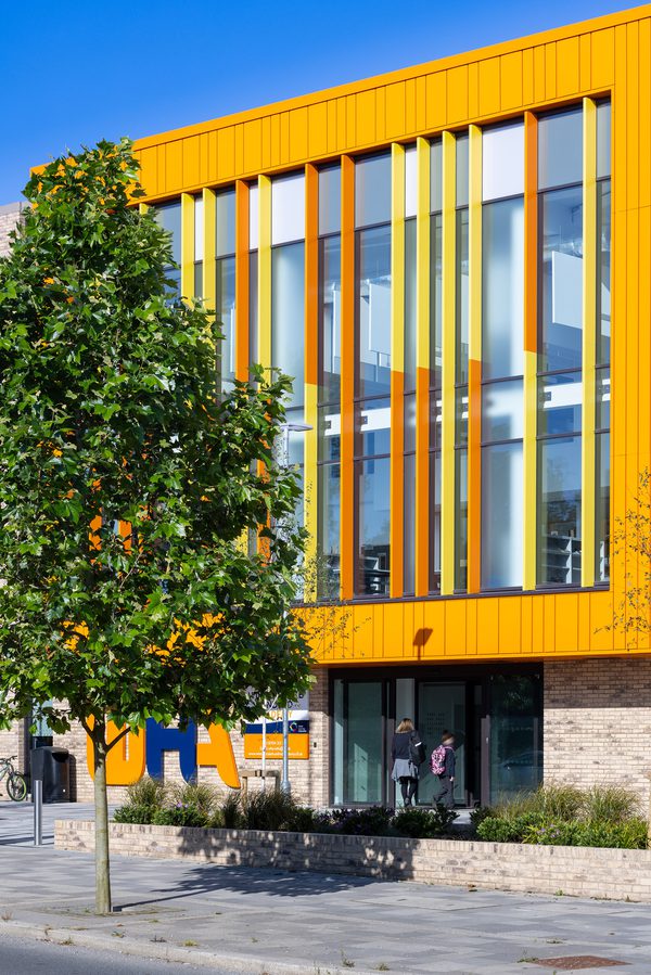 Students using the entrance of the Winterstoke Hundred Academy, with bright orange facade above and planting in front