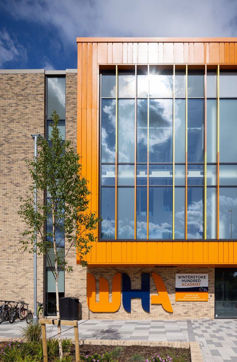 A detail shot of the front entrance of the Winterstoke Hundred Academy school, with high levels of glazing, orange facade, school logo and surrounding greenery.