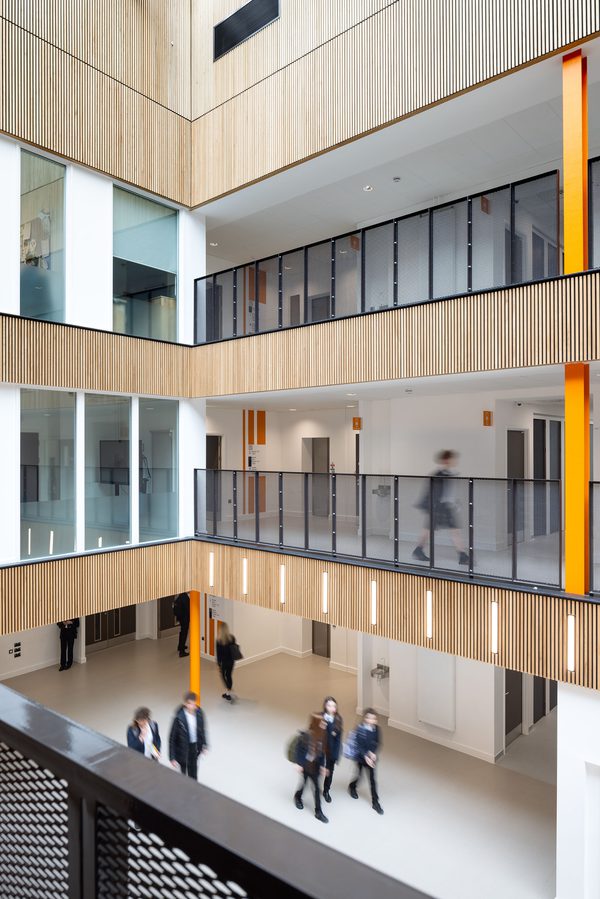 Students walking through the triple height, bright, central atrium within Winterstoke Hundred Academy, wooden facade detailing, glass fronted mezzanine levels and orange columns.